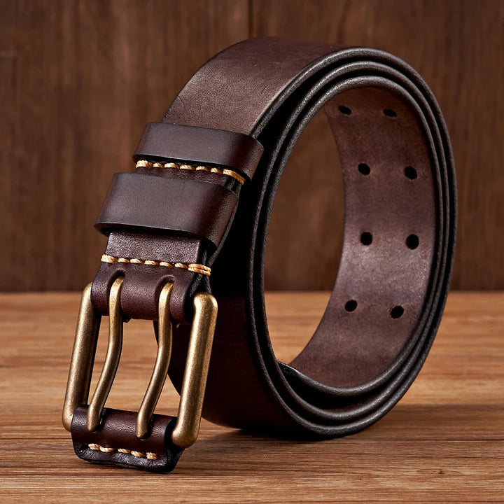 A rolled-up dark brown cowhide leather belt with a double copper buckle and contrast stitching, displayed on a wooden surface with a wood-paneled background.