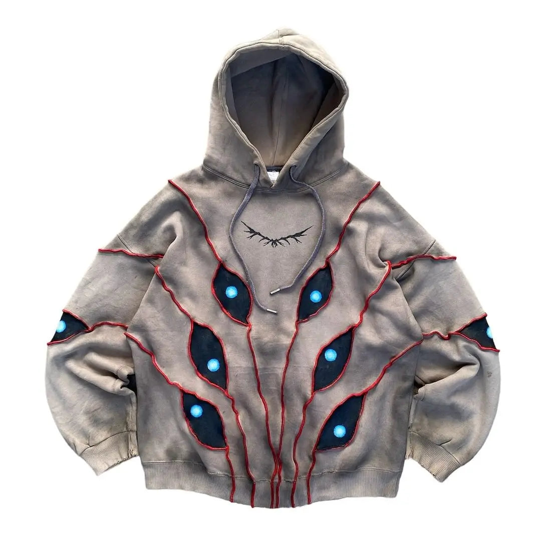 A gray hoodie featuring red stitched lines and black shapes with blue circles in an eye pattern patchwork, radiating from the center. The chest area has a curved stitched line like a closed eye, made with breathable fabric for comfort.