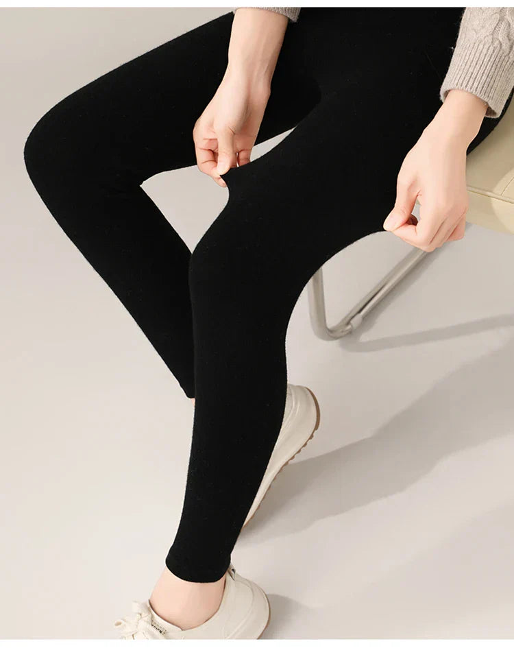 A person wearing black high-waisted leggings, a beige sweater, and white shoes is seated on a chair. The person is pulling at the material of the leggings to show their stretchiness.