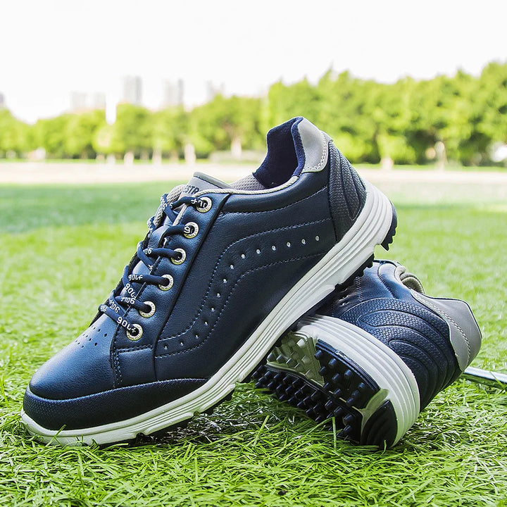 A pair of navy blue golf footwear with white soles, featuring advanced cleat technology, placed on green grass with trees and city buildings blurred in the background.