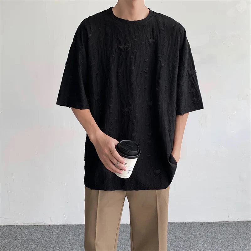 A person wearing an oversized black textured streetwear tee and beige pants stands against a plain light-colored wall, holding a white takeaway coffee cup in one hand with the other hand in their pocket.