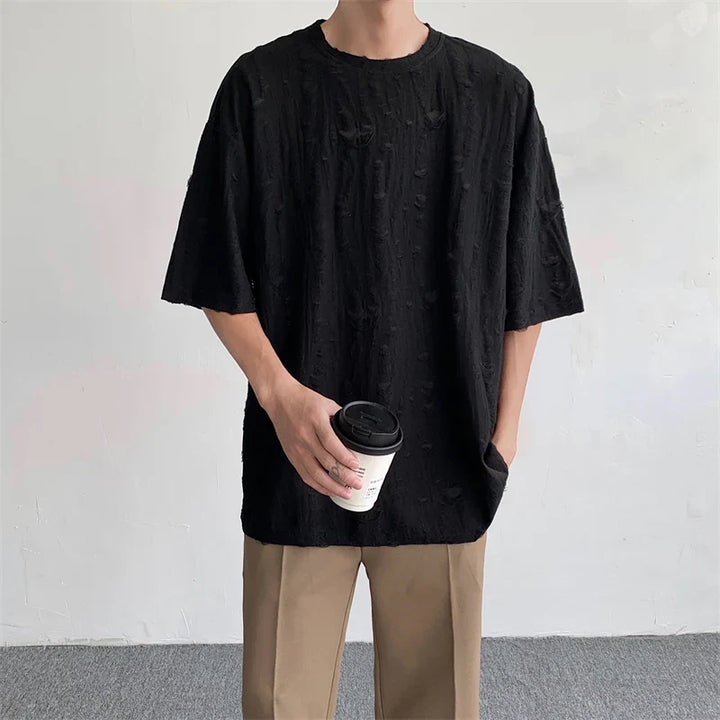 A person wearing an oversized black textured streetwear tee and beige pants stands against a plain light-colored wall, holding a white takeaway coffee cup in one hand with the other hand in their pocket.