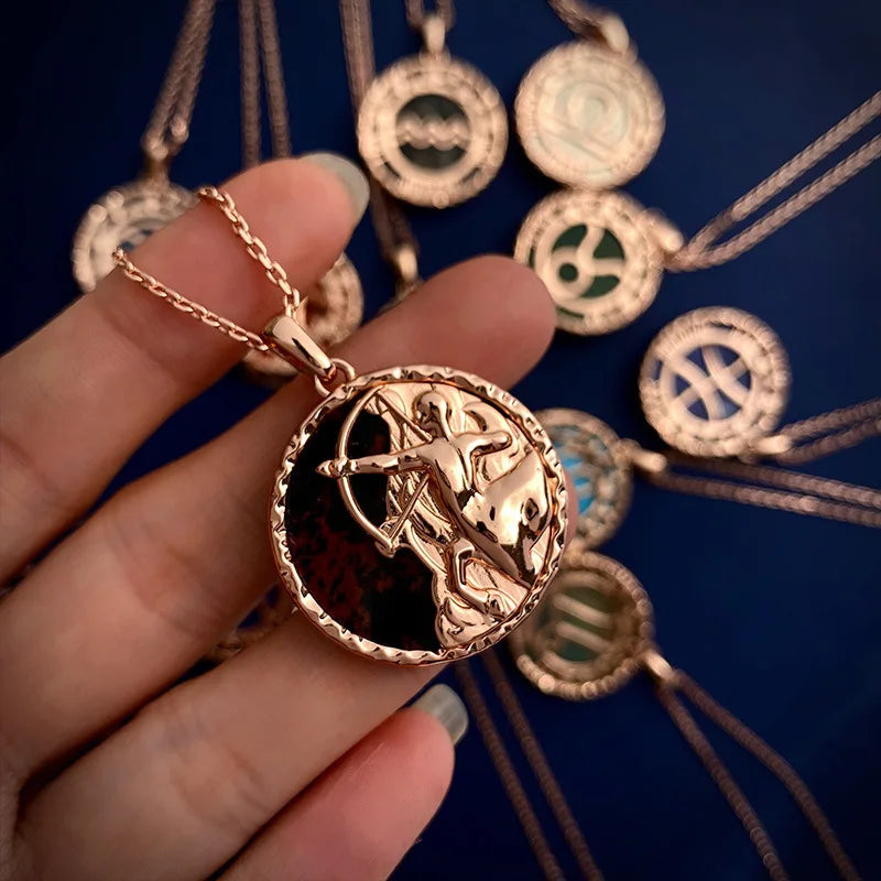 A hand holds a rose gold zodiac necklace featuring a Capricorn symbol; similar astrology jewelry pendants with a copper finish are arranged in the background on a dark blue surface.