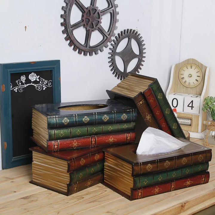 A stack of decorative tissue boxes designed to look like vintage book tissue boxes sits on a wooden desk, with gears, a small chalkboard, and a clock in the background. One tissue partially emerges from a book-shaped box.