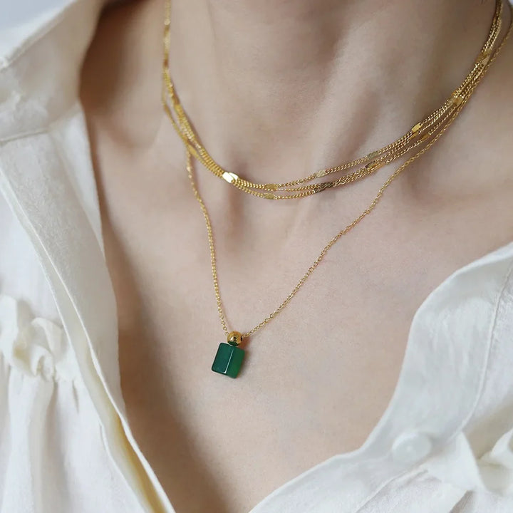 A person wearing a white shirt is shown wearing layered 18K gold plated necklaces, including a short multi-strand chain and a longer chain with a rectangular green zirconia pendant.