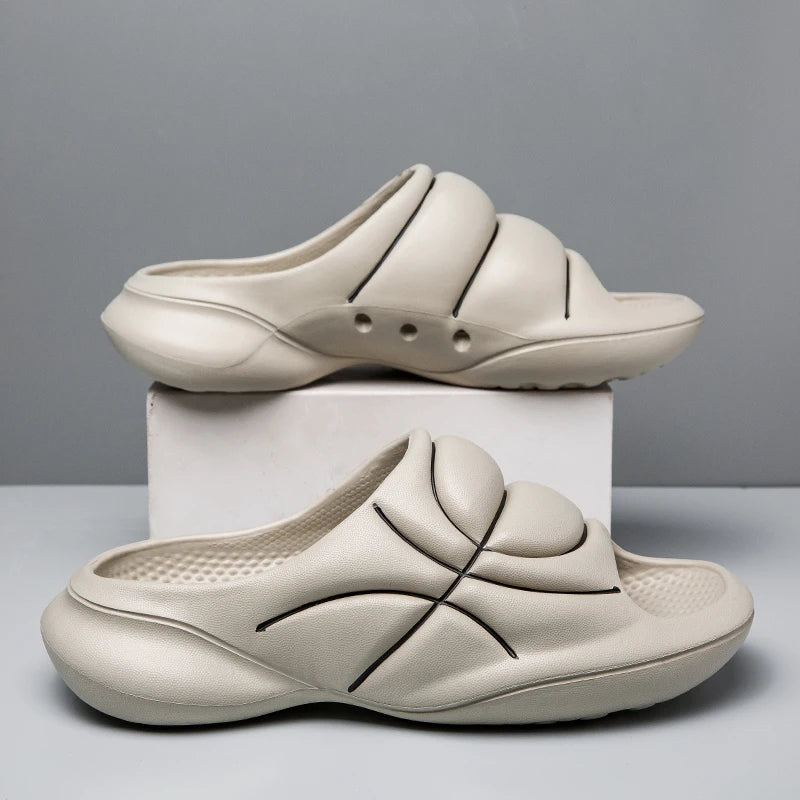 Two beige slide sandals with a cushioned footbed and chunky design feature black curved line accents, one resting on a white box and the other on the table, against a plain gray background.