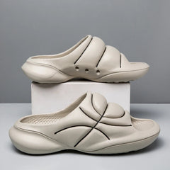 Two beige slide sandals with a cushioned footbed and chunky design feature black curved line accents, one resting on a white box and the other on the table, against a plain gray background.