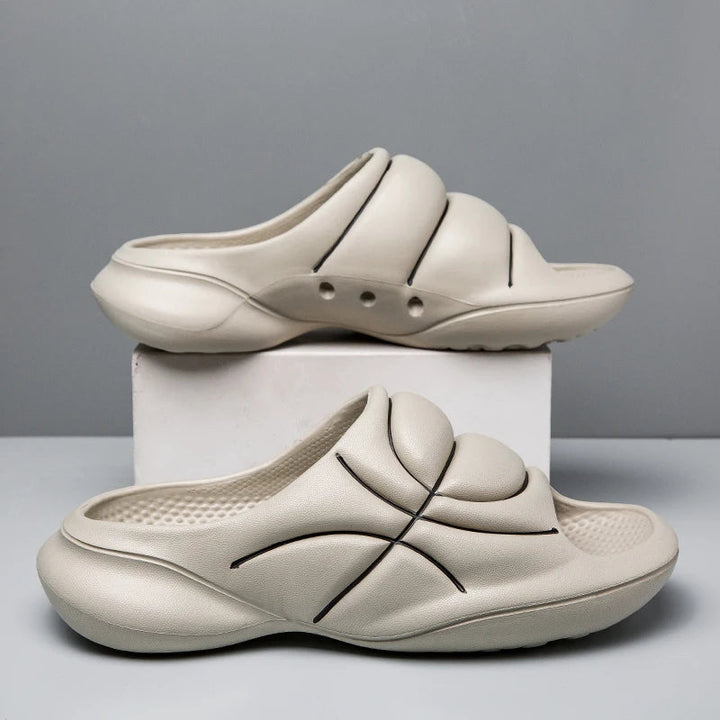 Two beige slide sandals with a cushioned footbed and chunky design feature black curved line accents, one resting on a white box and the other on the table, against a plain gray background.