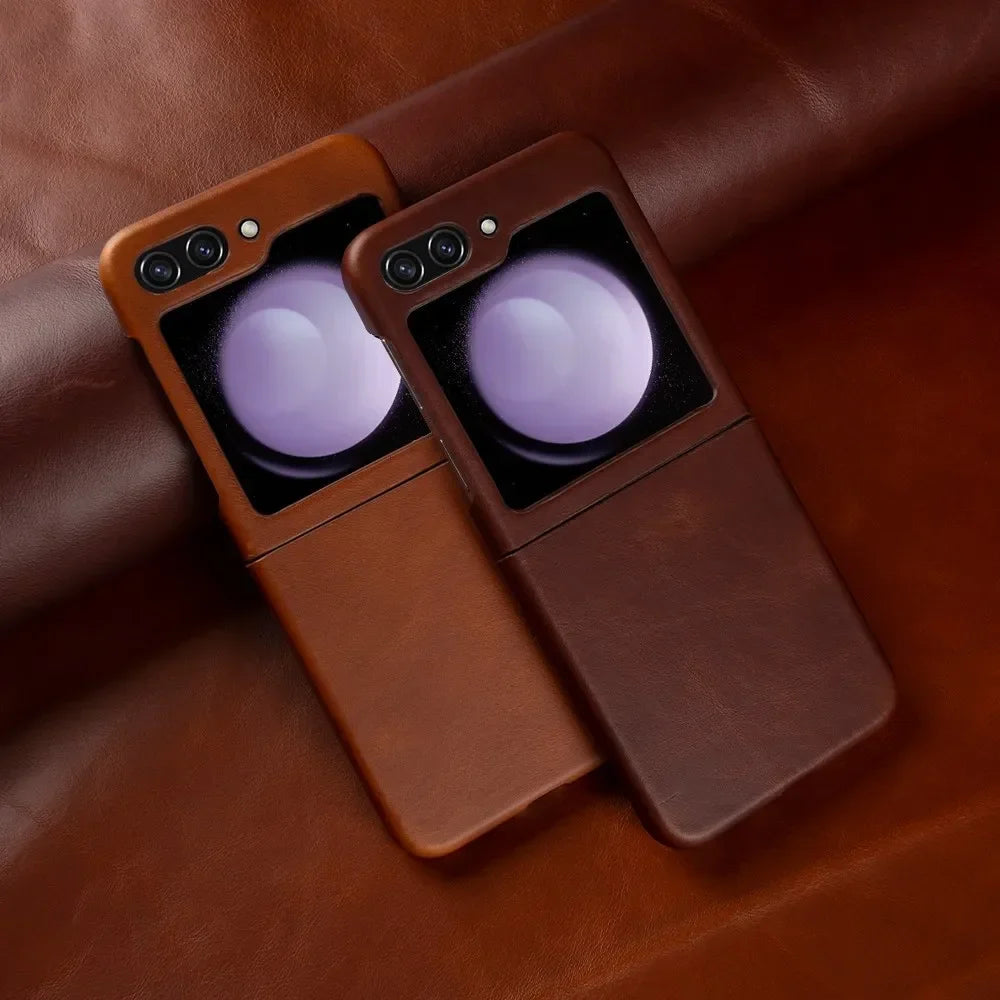 Two smartphones, possibly Samsung Galaxy Z Flip models, in brown genuine leather cases rest on a matching brown leather surface. Both cases offer shockproof protection and feature dual cameras with a reflective, circular accent near the camera area.