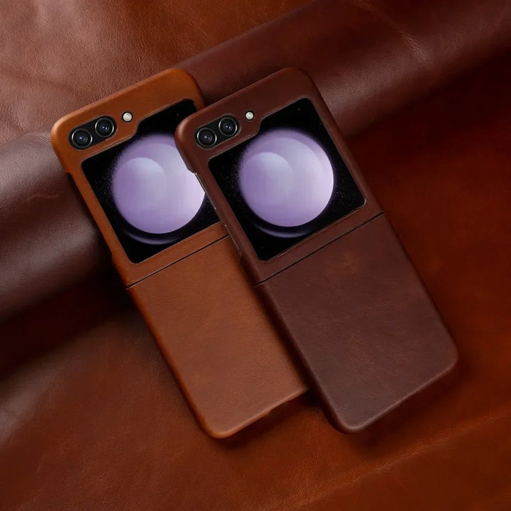 Two smartphones, possibly Samsung Galaxy Z Flip models, in brown genuine leather cases rest on a matching brown leather surface. Both cases offer shockproof protection and feature dual cameras with a reflective, circular accent near the camera area.