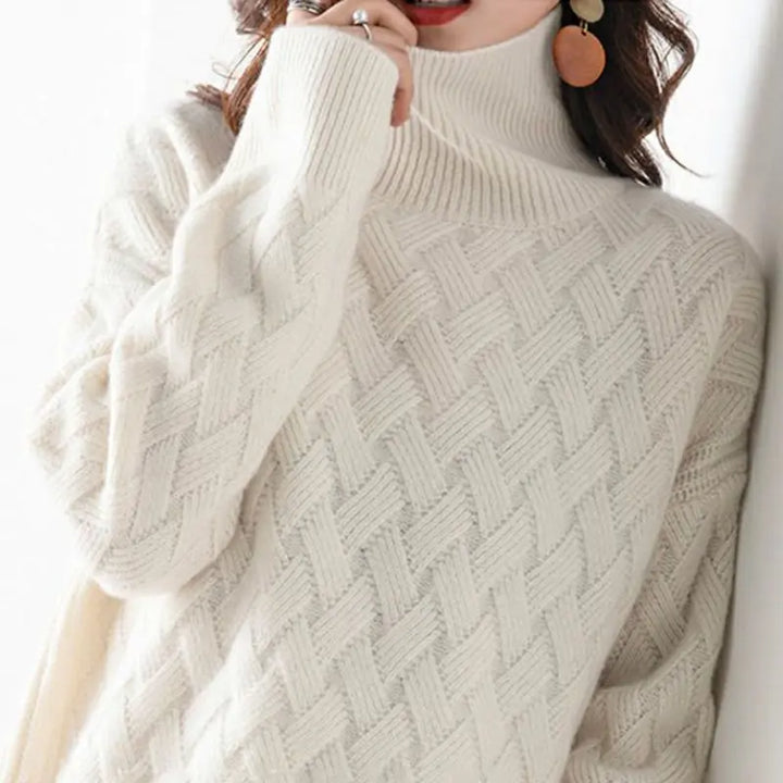 A person wearing a white, ultra soft, high-neck knit sweater with a basketweave pattern. They have wavy hair, large round earrings, and are partly covering their mouth with the sweater sleeve.