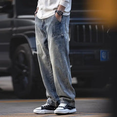 A person wearing loose-fitting, classic denim blue jeans, a white long-sleeve shirt, a watch, and black-and-white sneakers stands outdoors with hands in pockets, in front of a dark-colored vehicle.