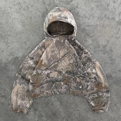 A camo pattern hoodie with a front pocket and hood, laid flat on a gray, textured concrete surface. Crafted in earthy tones of brown, green, and beige from a soft cotton blend for comfort.