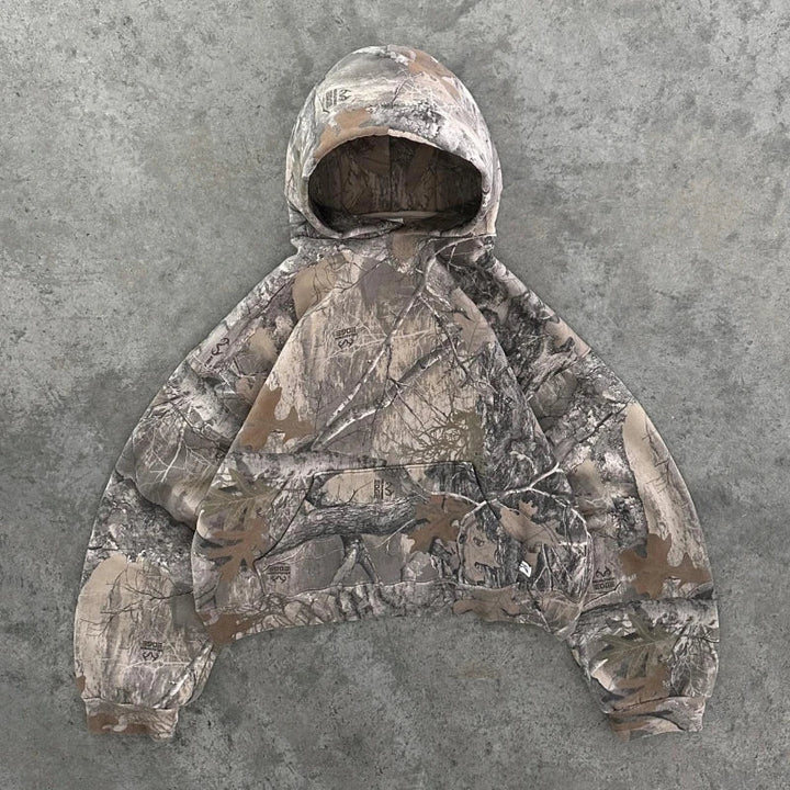 A camo pattern hoodie with a front pocket and hood, laid flat on a gray, textured concrete surface. Crafted in earthy tones of brown, green, and beige from a soft cotton blend for comfort.