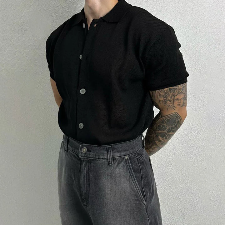 A person stands against a light wall, hands behind their back, wearing a short-sleeve unisex top styled as a black button-up and high-waisted gray jeans, with tattooed arms adding edge to the look.
