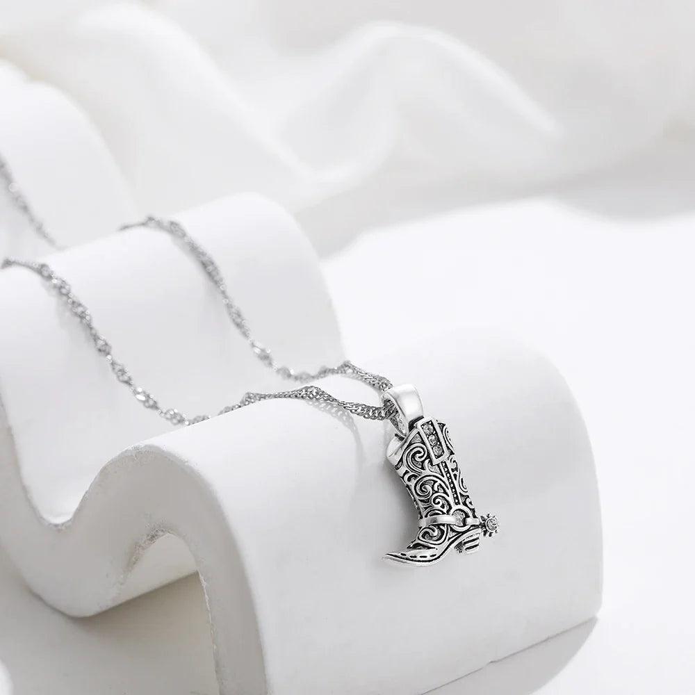 Silver cowgirl necklace featuring a detailed cowboy boot pendant, elegantly displayed on a white curved stand against a soft white fabric background—a timeless piece of western jewelry.