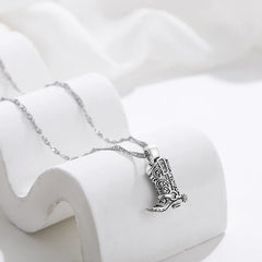Silver cowgirl necklace featuring a detailed cowboy boot pendant, elegantly displayed on a white curved stand against a soft white fabric background—a timeless piece of western jewelry.