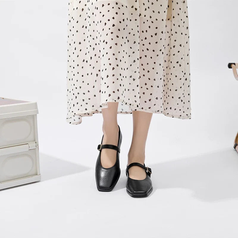 A person wearing a white midi skirt with black heart patterns and Augusta Leather Ballet Flats stands on a white floor near a white chest and a beige chair.