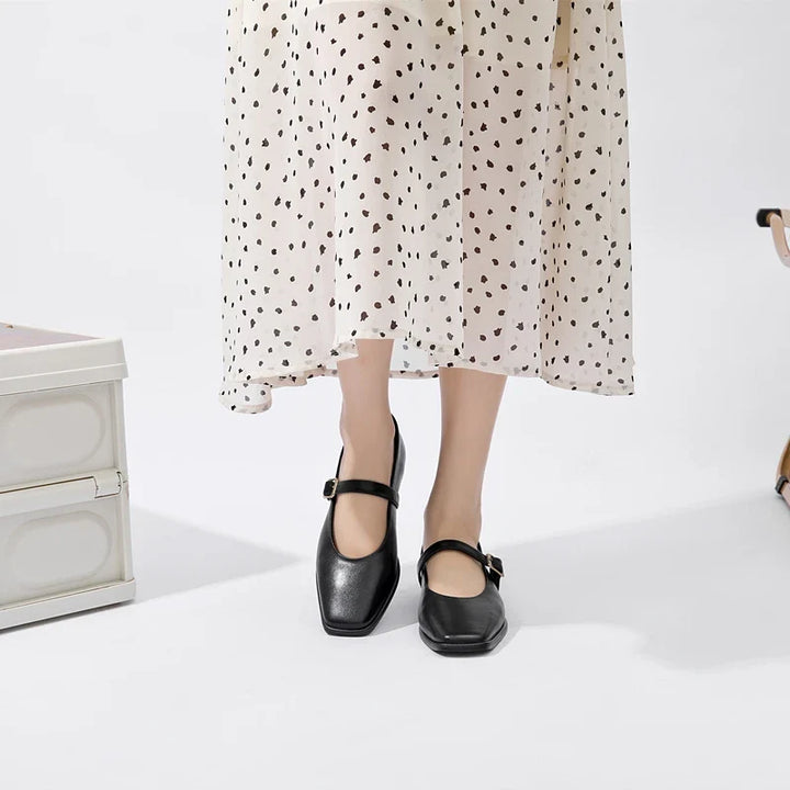 A person wearing a white midi skirt with black heart patterns and Augusta Leather Ballet Flats stands on a white floor near a white chest and a beige chair.