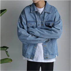 Person standing with arms crossed, wearing an oversized light blue premium denim jacket over a white T-shirt and black pants, with a neutral gray background and a plant partially visible on the left side.