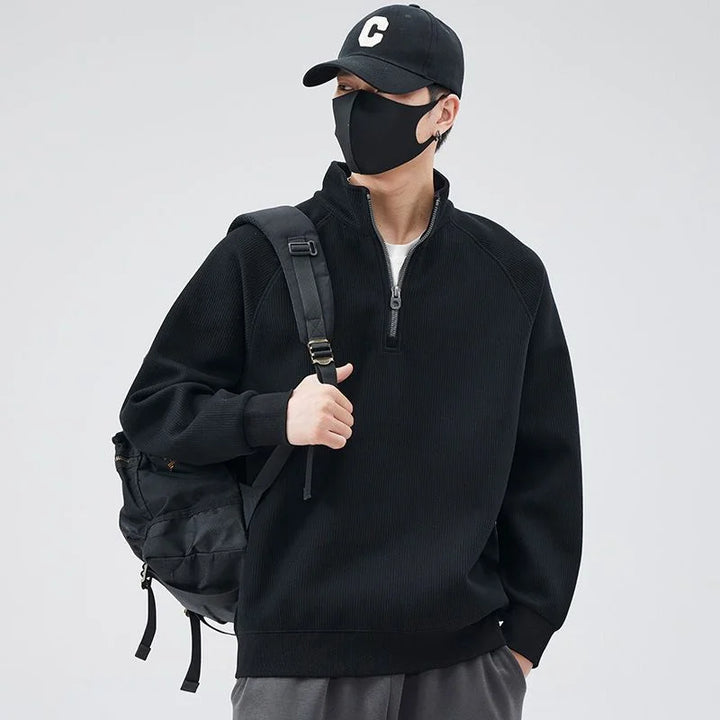 A person in a black cap with a C, black face mask, relaxed fit black half-zip sweater with stand-up collar, and a black backpack stands against a plain light background, looking to the side.