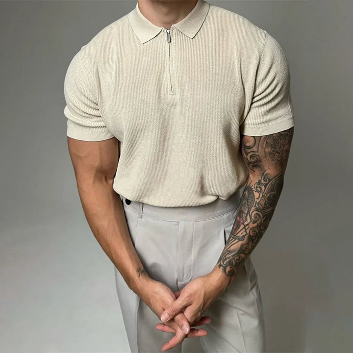 A person with muscular arms and a tattooed forearm wears a light beige viscose polo with ribbed detailing, tucked into matching beige pants, standing against a plain background with hands clasped in front.