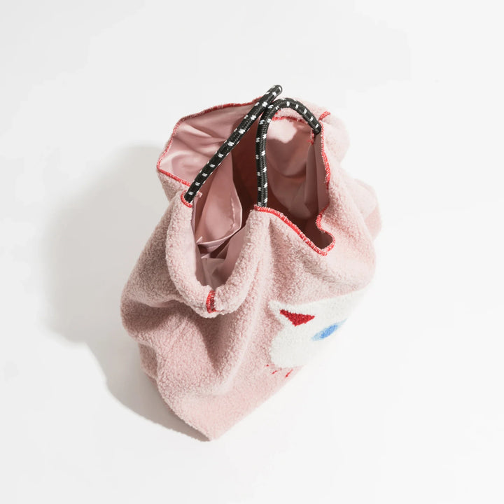 A pink drawstring faux fur handbag with a fuzzy, lamb-like fabric, black and white rope handles, red trim, and a partially visible colorful design on the side, photographed from above on a white background.