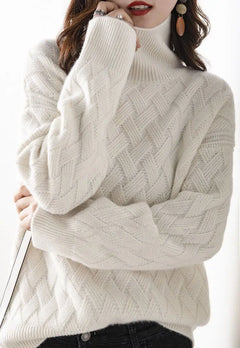 Woman wearing a white, ultra soft cable-knit turtleneck sweater with long sleeves, black pants, statement earrings, and red lipstick, posing with one hand near her face against a white background.
