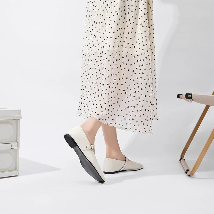 A person wearing a white polka dot dress and Augusta Leather Ballet Flats stands near a beige folding chair and a white storage box in a minimal, bright room.