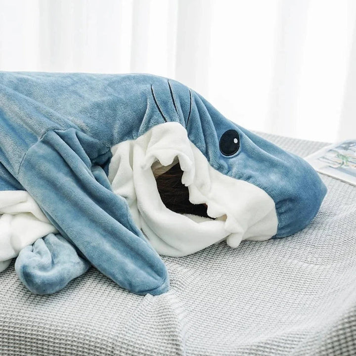 A person is lying on a bed, mostly covered by a large, plush shark sleeping bag, with just their face partially visible inside the shark’s open mouth. The background is bright and softly lit.