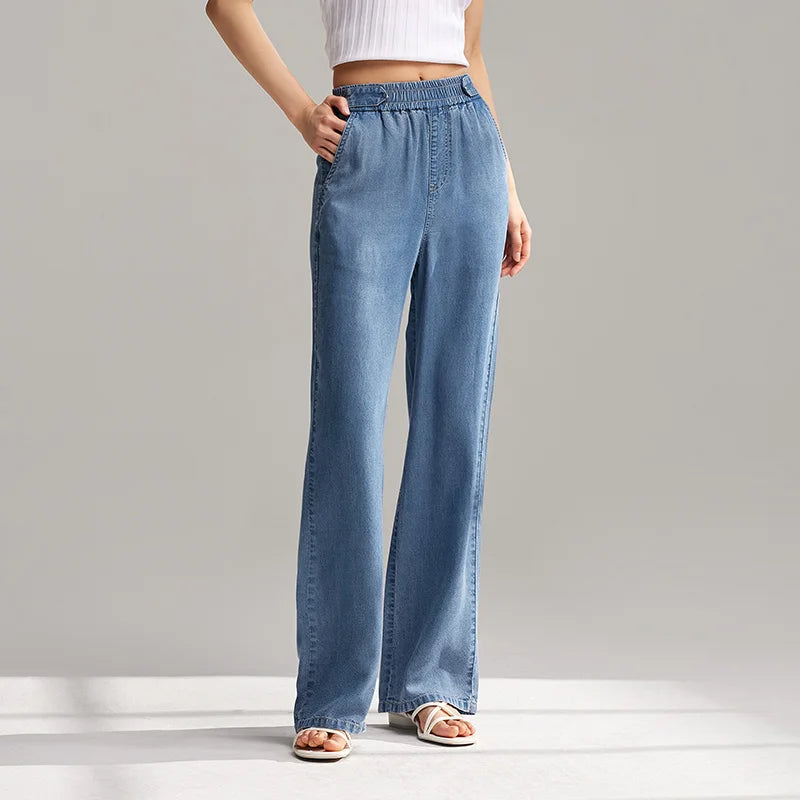 A person wearing light blue, breathable denim wide-leg jeans with an elastic waistband, a white sleeveless crop top, and white sandals stands against a plain gray background.