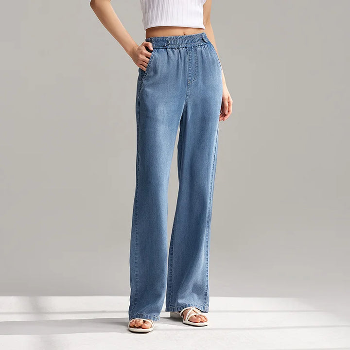 A person wearing light blue, breathable denim wide-leg jeans with an elastic waistband, a white sleeveless crop top, and white sandals stands against a plain gray background.