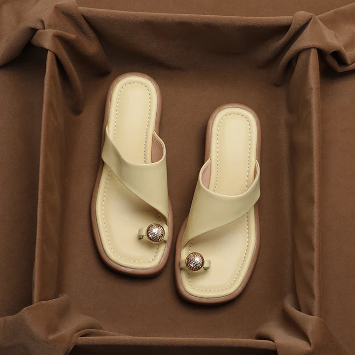 A pair of beige Square Toe Slides with a toe loop and decorative gold and brown beads, displayed on a soft brown fabric background—perfect for those seeking Women's Modern Slippers with a fashionable touch.