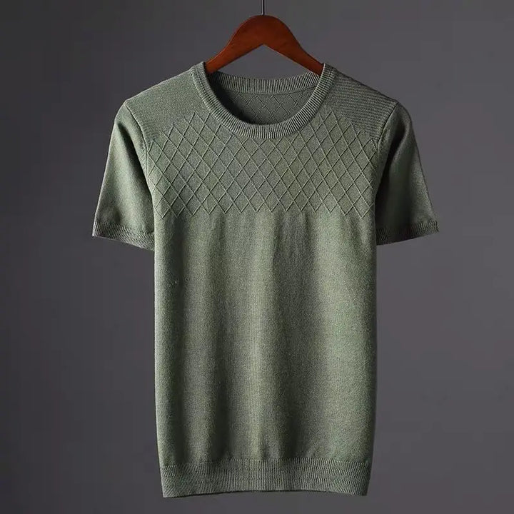A short-sleeved, olive green knit silk-cotton tee with a classic fit and a diamond-pattern across the upper chest, hanging on a wooden hanger against a plain gray background.