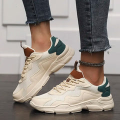 A person wearing beige chunky slip-on street shoes with green accents and brown details, featuring advanced shock absorption, paired with frayed-edge skinny jeans and a silver anklet, standing on a dark wooden floor against a white textured wall.