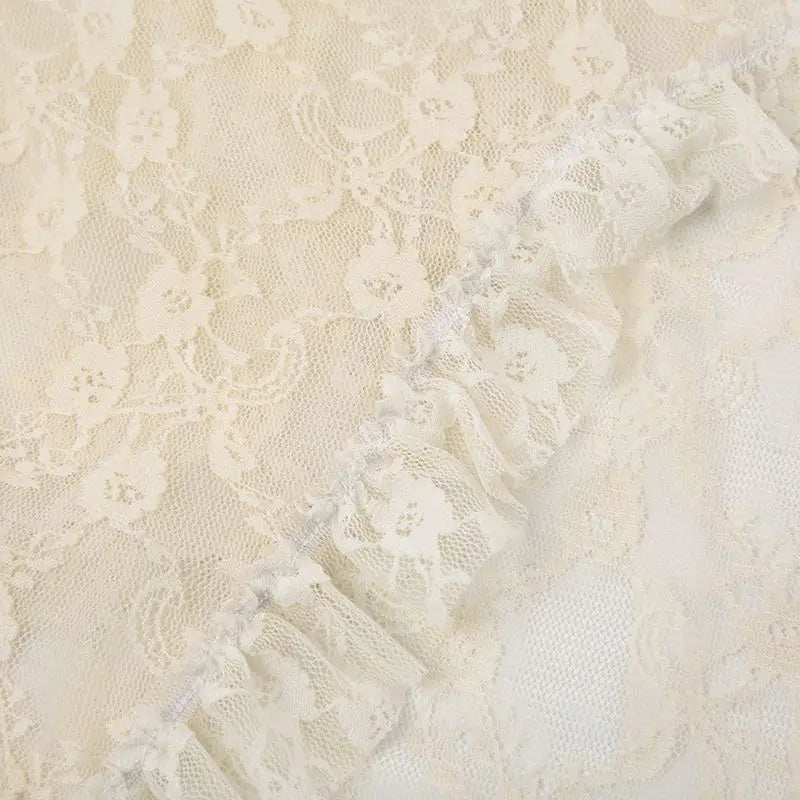 Close-up of cream-colored silky lace fabric with delicate patterns and a gathered ruffled edge running diagonally across the image, evoking an asymmetrical hemline.