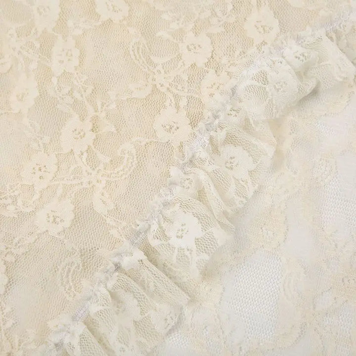 Close-up of cream-colored silky lace fabric with delicate patterns and a gathered ruffled edge running diagonally across the image, evoking an asymmetrical hemline.