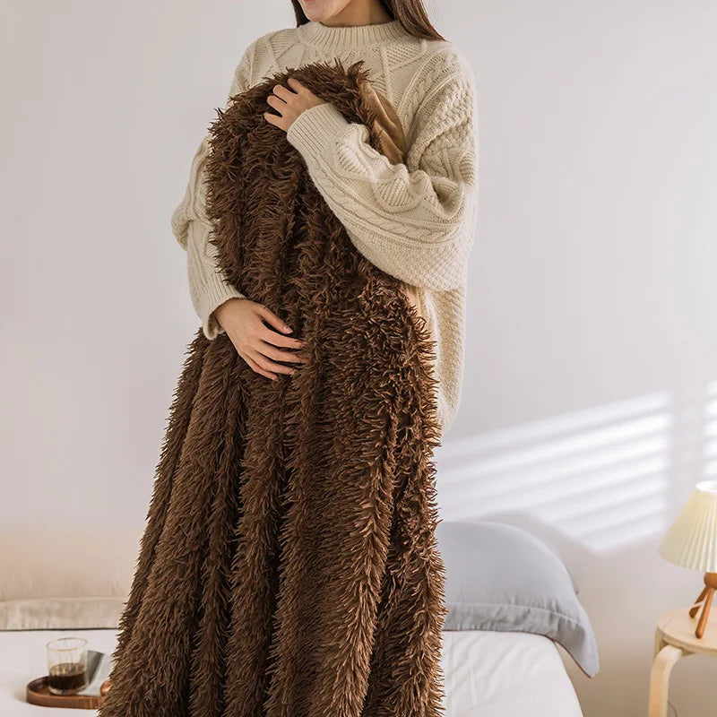A person in a cream-colored sweater stands in a warmly lit bedroom, holding a large, fluffy Soft Faux Fur Throw Blanket. The bed, side table with a lamp, and glass of water add to the warm and cozy ambiance of the space.