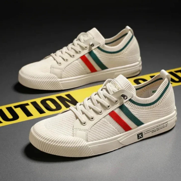 A pair of white canvas sneakers with red and green stripes on the sides, featuring a durable rubber outsole, placed on a dark surface near yellow caution tape.