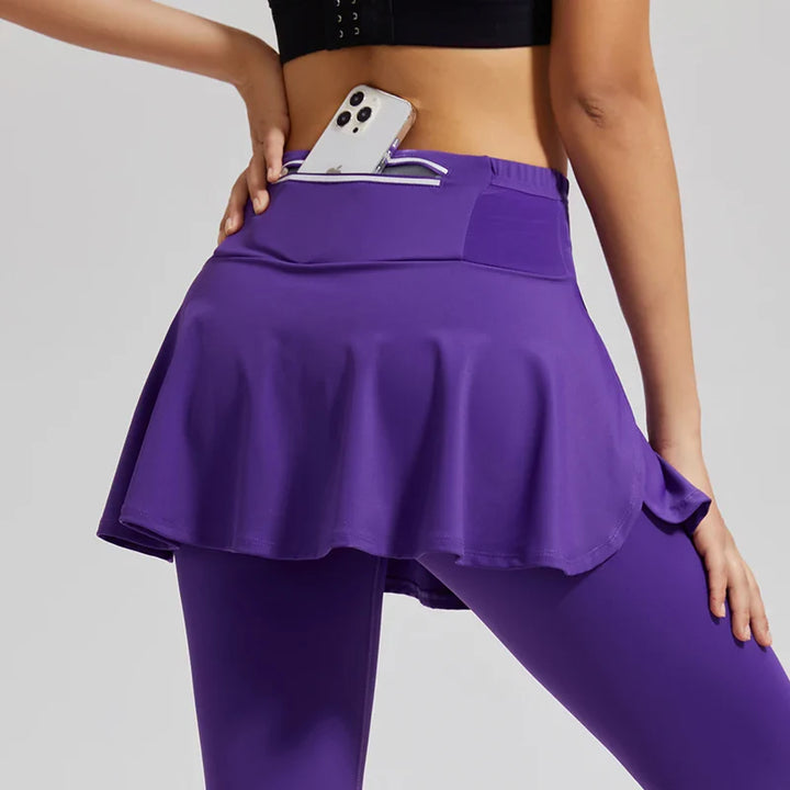 A person wearing black skirted leggings with high-waisted support and a purple athletic skirt stands with a hand on their hip. A smartphone is partially tucked into a stretch storage pocket on the waistband of the skirt.