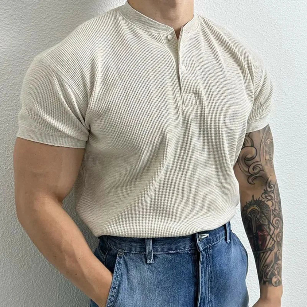 A person wearing a short-sleeved, cream-colored ribbed Henley and blue jeans stands against a white wall, showing a muscular build and a detailed tattoo on their left forearm.