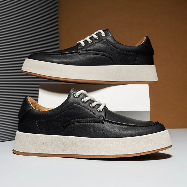 A pair of black leather sneakers, inspired by Italian designer shoes, with white laces and thick white soles are displayed stylishly—one balanced on a white box, the other resting on a flat surface.