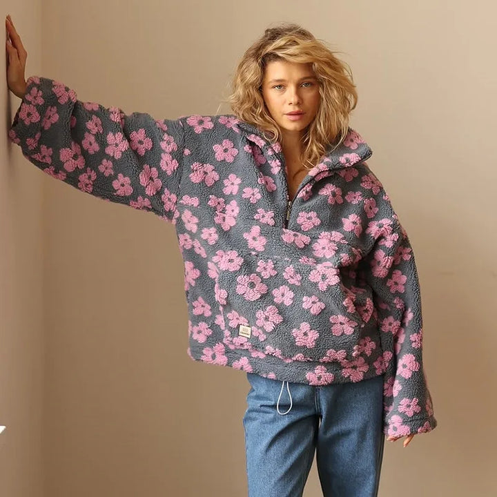 A woman with wavy blonde hair wears a loose, grey flannel pullover with a pink floral pattern, blue jeans, and stands against a plain beige wall with one arm resting on the wall.