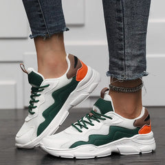 A person wearing lightweight mesh slip-on street shoes with green, brown, and orange accents, paired with frayed-hem skinny jeans, stands on a dark floor in front of a white wall.