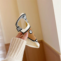 A hand in a beige sweater holds a soft silicone phone case shaped like a cartoon dog with a raised ear, tan face, and big white eyes, offering shockproof protection in a softly lit room with light brown and white walls.