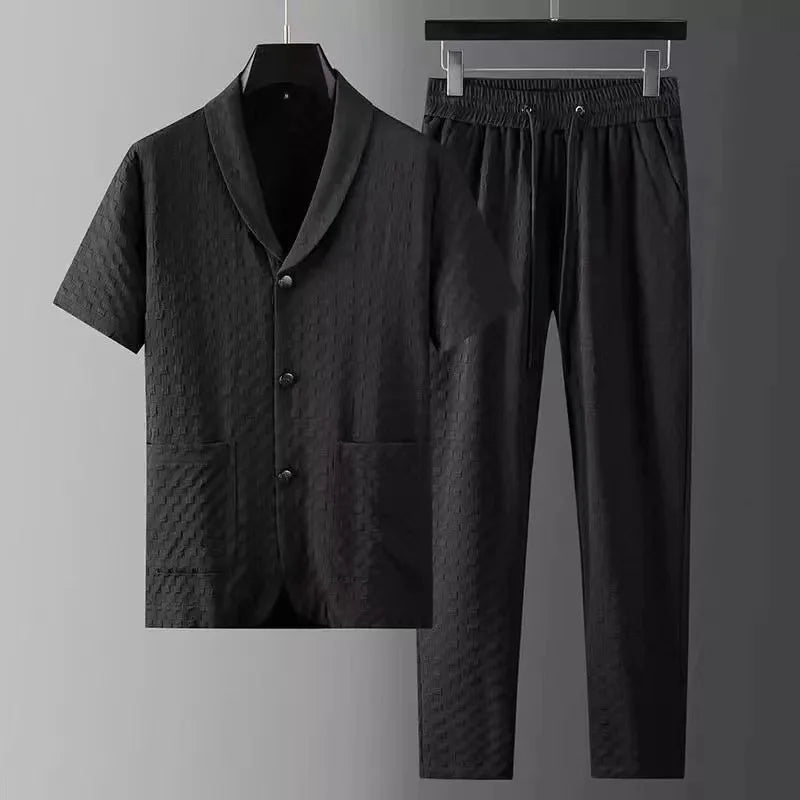 A matching black two-piece set featuring a short-sleeve button-up shirt with notch lapel and two front pockets, paired with black pants featuring an elastic drawstring waistband, both with a subtle textured pattern, displayed on black hangers.