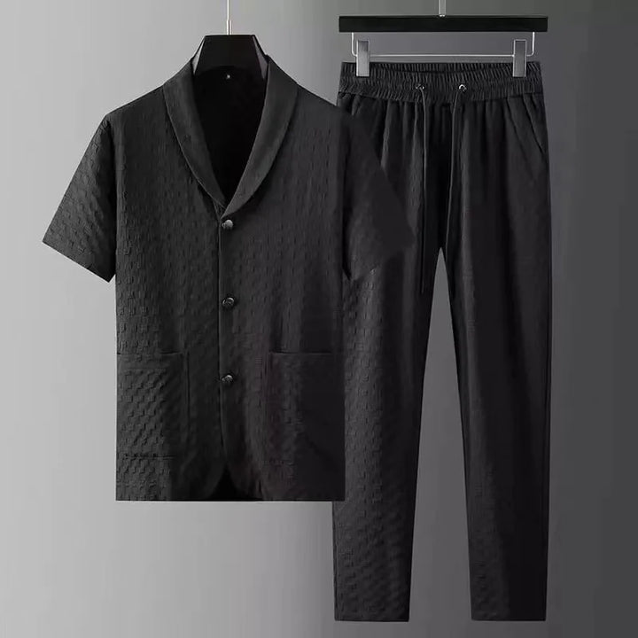 A matching black two-piece set featuring a short-sleeve button-up shirt with notch lapel and two front pockets, paired with black pants featuring an elastic drawstring waistband, both with a subtle textured pattern, displayed on black hangers.