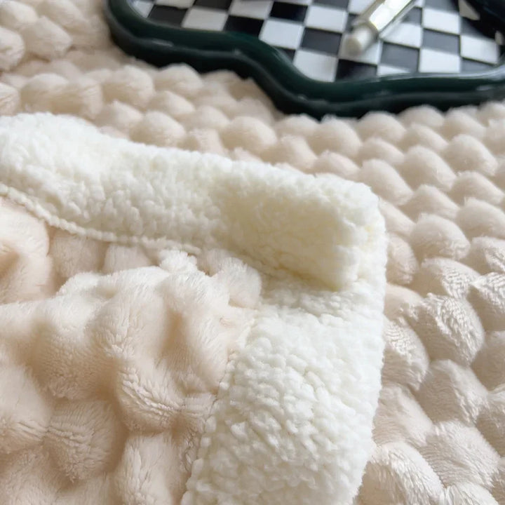 A close-up of a textured beige and white ultra-soft cotton blanket with a fluffy trim, lying on a soft surface. In the background, part of a checkerboard-patterned tray and a pen are visible.