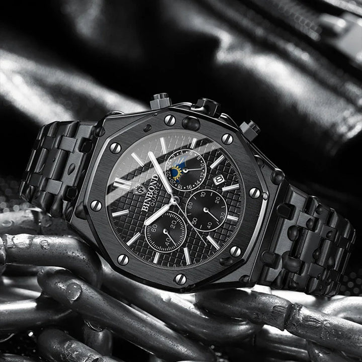 A black luxury stainless steel watch with a metal band and multiple chronograph dials is displayed on a background of metallic chains and dark leather. The watch face features silver accents, prominent screw details, and precision quartz movement.