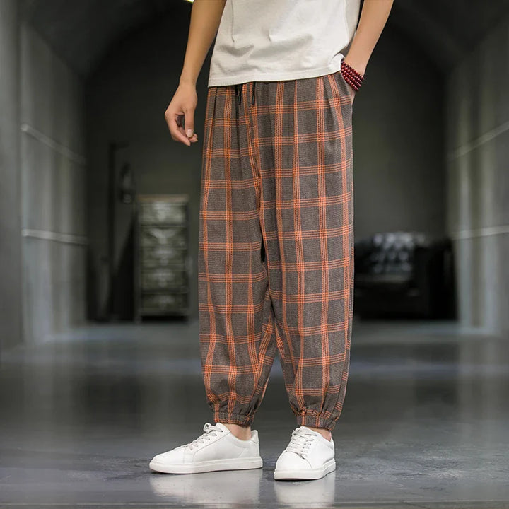 A person showcases Tokyo streetwear, wearing a white T-shirt and loose-fitting plaid pants with orange and gray patterns, standing indoors on a shiny floor with hands in pockets and white sneakers—a modern fashion statement.